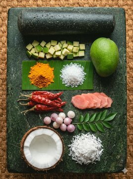 Traditional Sri Lankan raw mango chutney or curry ingredients laid out flat lay on a green granite grinding stone (miris gala) with a pestle, featuring green mango, coconut, dried red chillies, shallo
