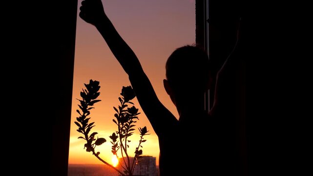 Peace silhouette of young child stretching arms after waking up in front of window. Kid enjoying beautiful warm sunrise over city and sea, symbolizing hope and a new day