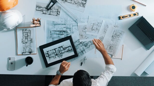 Top view of skilled engineer selecting material for house design while sitting at meeting table with tablet, laptop, safety helmet and equipment. Smart architect looking at project plan. Alimentation.