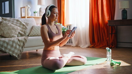 Fototapeta premium Sporty asian girl browsing phone before yoga meditation. Lotus pose home mat.