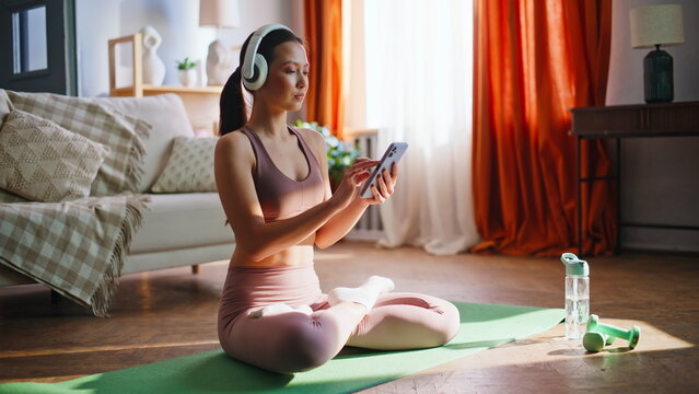 Sporty asian girl browsing phone before yoga meditation. Lotus pose home mat.