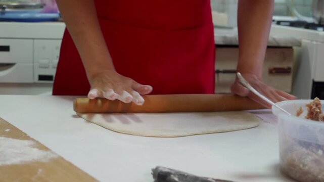 Close view of hands pressing dough with wooden rolling pin over flour dust on white countertop, with red apron in background and ingredient containers nearby, creating warm home baking atmosphere