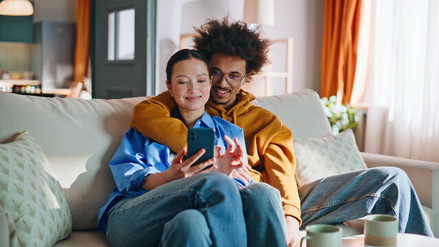 Multiethnic couple browsing smartphone together on sofa at home. Cozy weekend.