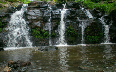 A scenic tropical waterfall cascading over dark volcanic rocks, surrounded by lush green bamboo and dense rainforest vegetation in Indonesia. Captured with a long exposure technique, the silky flowing © Eriec