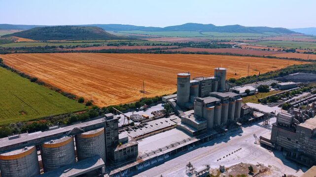 Concrete silos at an industrial facility. An aerial high angle perspective shows rows of large grey storage silos next to a vibrant golden field.