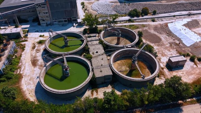 Circular industrial water treatment tanks from an aerial view. Two clarifiers contain green liquid while two remain dry inside a large factory compound on a sunny day.