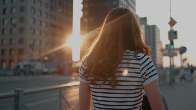 Back view of a young mother walking with a baby stroller on a city street during a stunning sunset. Woman strolling with her child in the evening, with golden sunlight creating a warm flare