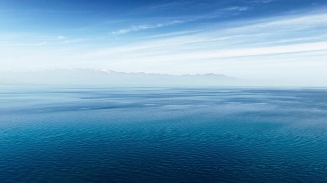 Aerial view of vast deep blue lake water stretching to distant misty mountain range under a clear sky.