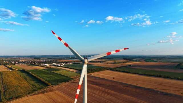 Wind turbine above geometric agricultural fields. Aerial view shows red marked blades over brown and green plots beside a compact village.