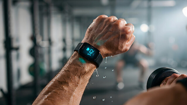 A sweaty muscular arm holding a black dumbbell in a gym, wearing a fitness tracker watch displaying a heart rate of 142 beats per minute on screen
