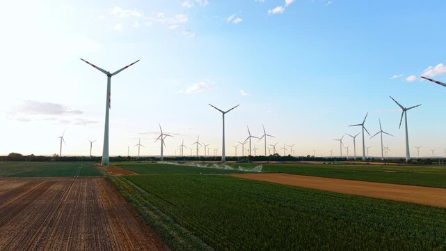 Wind farm across irrigated agricultural fields. Rows of turbines span the horizon while sprinklers spray green crops beneath a pale blue sky.
