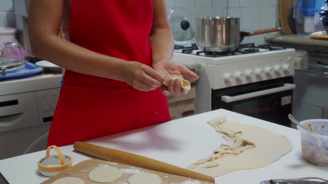 Close view of home baker using rolling pin to flatten dough on floured countertop, with pastry scraps and cookie cutters nearby, plus stovetop, saucepan, and utensils in warm kitchen background