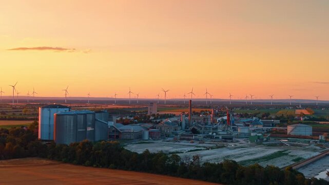 Industrial factory complex and wind turbine farm at sunset. Silos and factory buildings stand near fields under a vibrant orange sky during dusk.