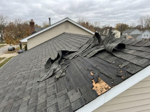 Damaged asphalt shingle roof after a storm, showing missing and torn shingles and exposed underlayment, with houses and trees in the background