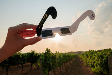 Obraz premium Hand holding certified solar eclipse viewing glasses against the bright sun in a vineyard landscape at sunset. The protective eclipse glasses are silhouetted by warm sunlight.