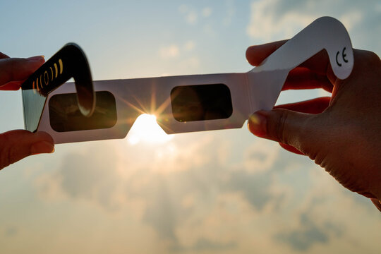 Hand holding certified solar eclipse viewing glasses against the bright sun in a vineyard landscape at sunset. The protective eclipse glasses are silhouetted by warm sunlight.
