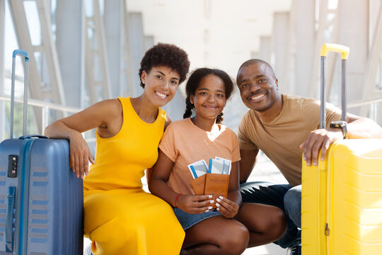 Happy Black family crouching together between rolling suitcases at bright airport terminal, smiling at the camera while their daughter holds passports and boarding passes