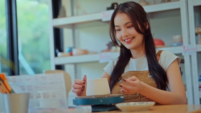 Closeup - Cheerful Asian woman proudly showing handmade ceramic mug in pottery studio, creative hobby and artistic lifestyle in modern craft workshop