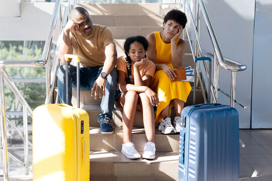 Tired African American family of three sitting on airport stairs, looking bored and exhausted while waiting near luggage suitcases, suffering flight delay