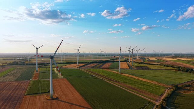 Wind farm turbine field during late afternoon. Long shadows stretch across colorful agricultural plots as multiple white structures stand under a bright sky.