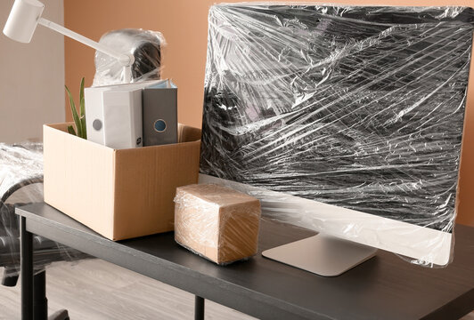 Office desk with computer monitor and cardboard boxes wrapped in plastic for moving or relocation, Transition of a workspace, with personal and professional items packed for transport
