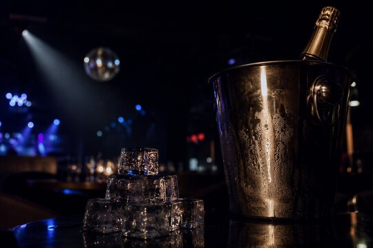 Champagne bottle in metal ice bucket with ice cubes on table. Night club atmosphere with disco ball and bokeh lights.