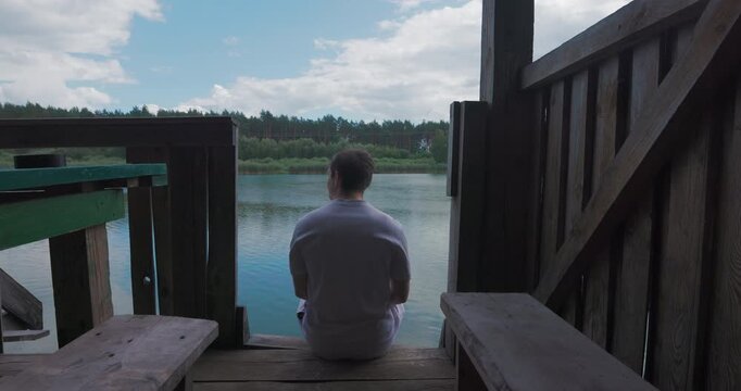 Young guy relaxing on the edge of wooden dock looking at river landscape