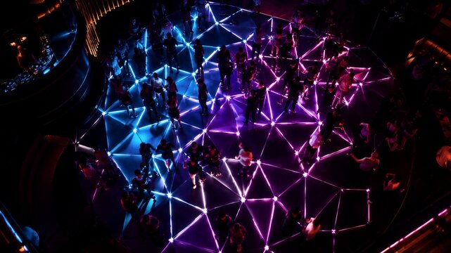 Crowd of people dancing on luminous geometric floor in nightclub. High angle view of party with neon triangle grid lighting.