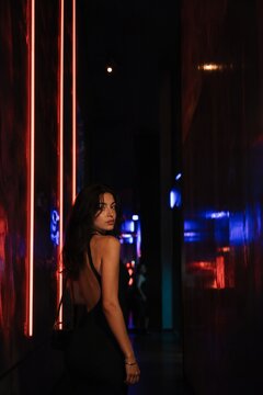 Woman in black dress posing in dark narrow corridor with red and blue neon lights. Cinematic portrait of stylish girl in night club. Urban nightlife, fashion and aesthetic mood.