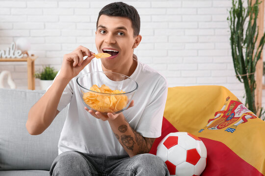 Young man with bowl of potato chips, soccer ball and flag of Spain watching football on sofa at home