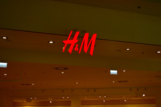 Illuminated red H&M store logo hanging from the ceiling inside a modern shopping mall center with warm interior lights.