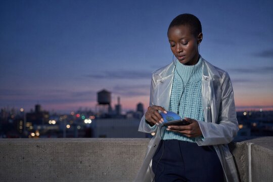 African american woman holding minidisc player on urban rooftop at twilight. Retro audio technology enthusiast listening to music. Concept of vintage media comeback and urban lifes