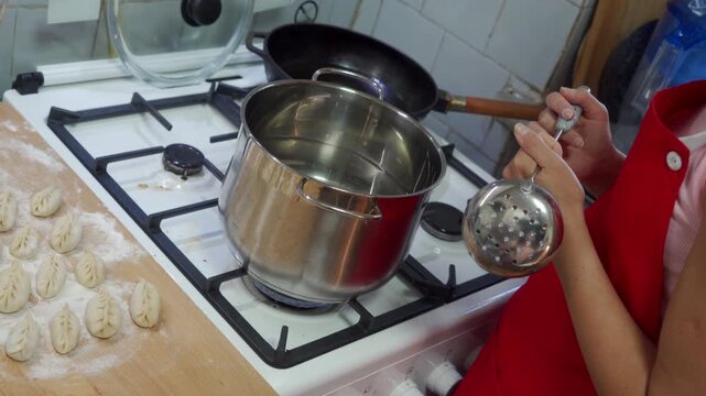 Close view of person wearing red apron ladling hot water from stainless steel pot on gas stove beside tray of raw dumplings; bright kitchen lighting, white tiled backsplash, metal ladle, stovetop