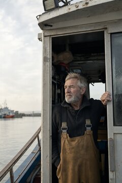 Fisherman standing in the doorway of a boat wheelhouse at the harbor. Mature man in work overalls looking at the horizon. Professional maritime industry and commercial fishing conc