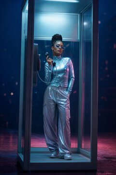 Woman in metallic clothing using old public phone inside glass booth at night. Futuristic portrait of fashionable girl with braids. Cyberpunk city style aesthetic and urban lifesty