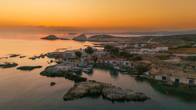 The fishing village of Mandrakia in Milos glows at sunrise, with colorful boathouses, calm Aegean waters, and soft golden light illuminating the cliffs and shoreline.