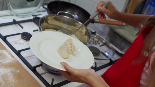 Close view of hands ladling thick creamy soup from stainless steel saucepan into shallow white bowl above lit gas stove, with flour dust, baking tools, countertop details, and warm domestic kitchen