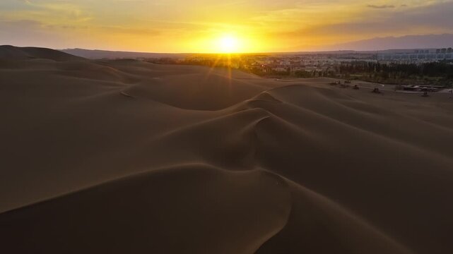 Aerial view of vast sand dune landscape in desert during a vibrant sunset near an oasis city.