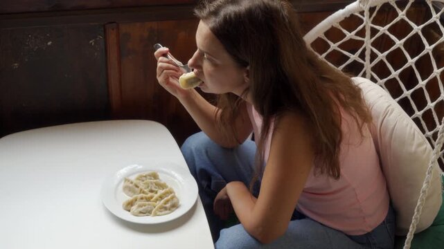 Casual young woman seated in wicker hanging chair beside bright window, lifting forkful of pasta from white bowl on small table; warm natural light, cozy home kitchen and soft neutral tones