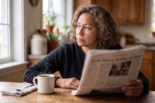 Mature diverse woman expresses political distrust and skepticism reading newspaper at kitchen table