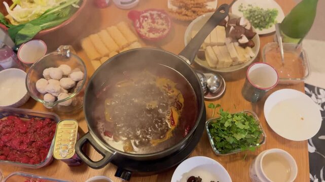 A cozy homemade hot pot feast served on a low dining table with a simmering soup base surrounded by thinly sliced beef, tofu, mushrooms, leafy vegetables, meatballs, dipping sauces, noodles, herbs