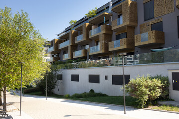 Fototapeta premium Modern residential building with balconies and decorative facade, surrounded by landscaped greenery and paved walkway in a contemporary urban setting