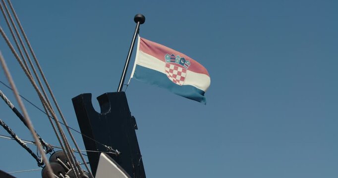 The Croatian national flag flies from the mast of a historic wooden sailing ship against a clear blue sky.