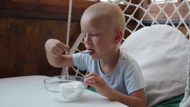 Blond boy sitting at white kitchen table stirring creamy yogurt in glass bowl with spoon while concentrating, natural window light softens pale skin, short hair, wood paneled wall, calm home