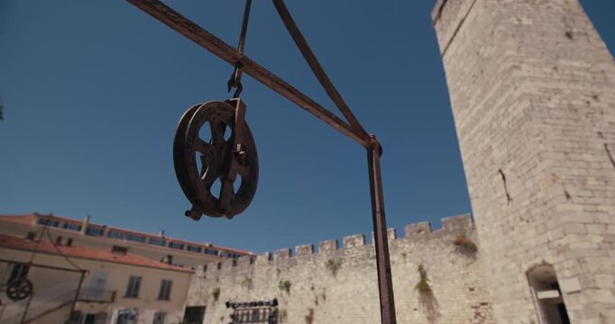 A historic iron well pulley stands in the foreground with the Captain Tower and medieval city walls in Five Wells Square in Zadar Croatia.