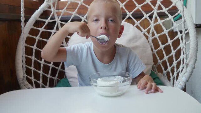 Blond toddler sits in round woven hanging chair and scoops white yogurt from glass bowl with spoon, surrounded by soft daylight, white table, cozy home setting, candid mealtime mood