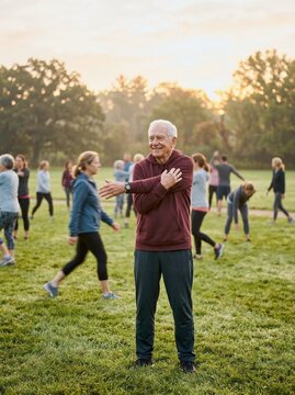 Senior man stretches on grass in park during sunrise as group exercises in warm light