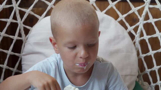 Blond young child sits in round woven hanging chair, eating melting ice cream with spoon, relaxed face and creamy mouth, soft natural light, neutral cushion background, cozy indoor mood