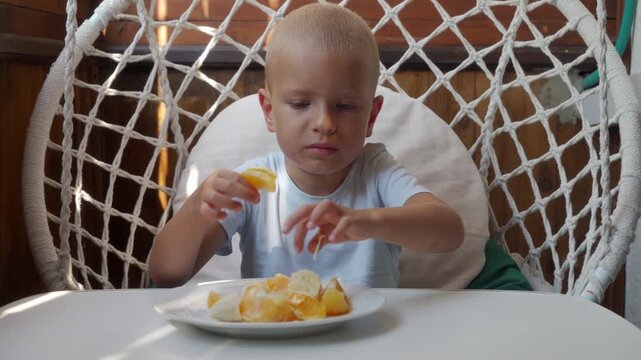 Toddler seated in white woven hanging chair beside round kitchen table peeling segmented orange with plate nearby, calm focused expression, soft window light, bright home dining scene