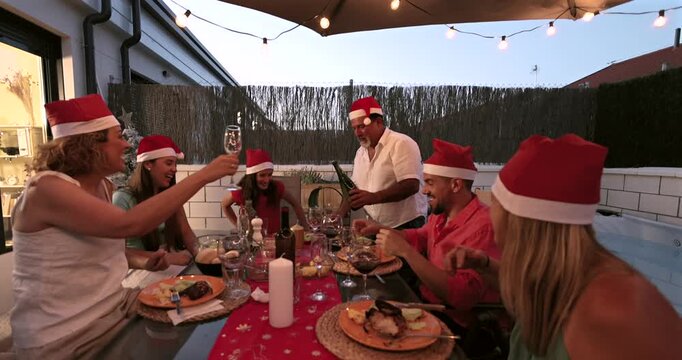 Cheerful family group wearing santa hats celebrating christmas with a festive dinner on an illuminated patio, with a man joyfully popping open and pouring a bottle of sparkling champagne